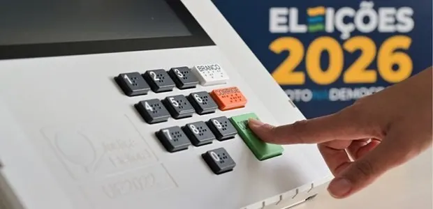 Dedo de eleitor pressiona a tecla verde Confirma de uma urna eletrônica; ao fundo, painel com o texto Eleições 2026