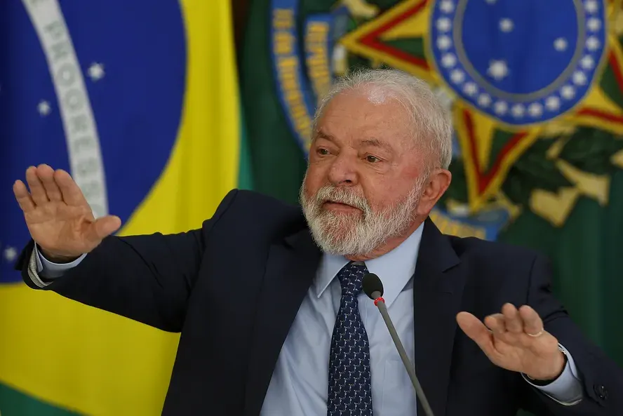 O presidente Luiz Inácio Lula da Silva, de cabelos e barba brancos, terno escuro e gravata azul, gesticula com as mãos abertas durante discurso no Palácio do Planalto; ao fundo, a bandeira do Brasil e o brasão da República